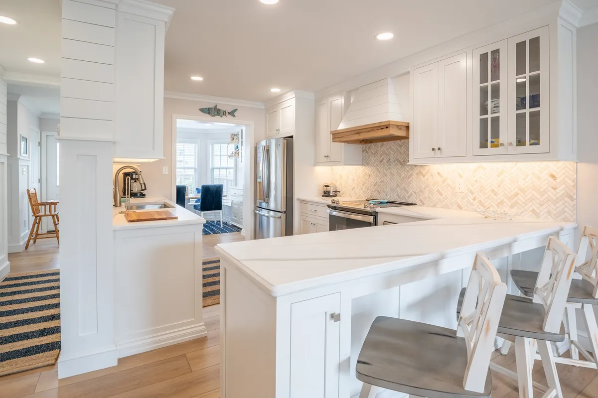 A bright, modern kitchen featuring a large island bar with countertop seating, gleaming stainless steel appliances, a stylish herringbone backsplash, and a striking wooden range hood. The space exudes friendliness and relaxation, perfect for cooking, entertaining, and enjoying coffee in the morning.