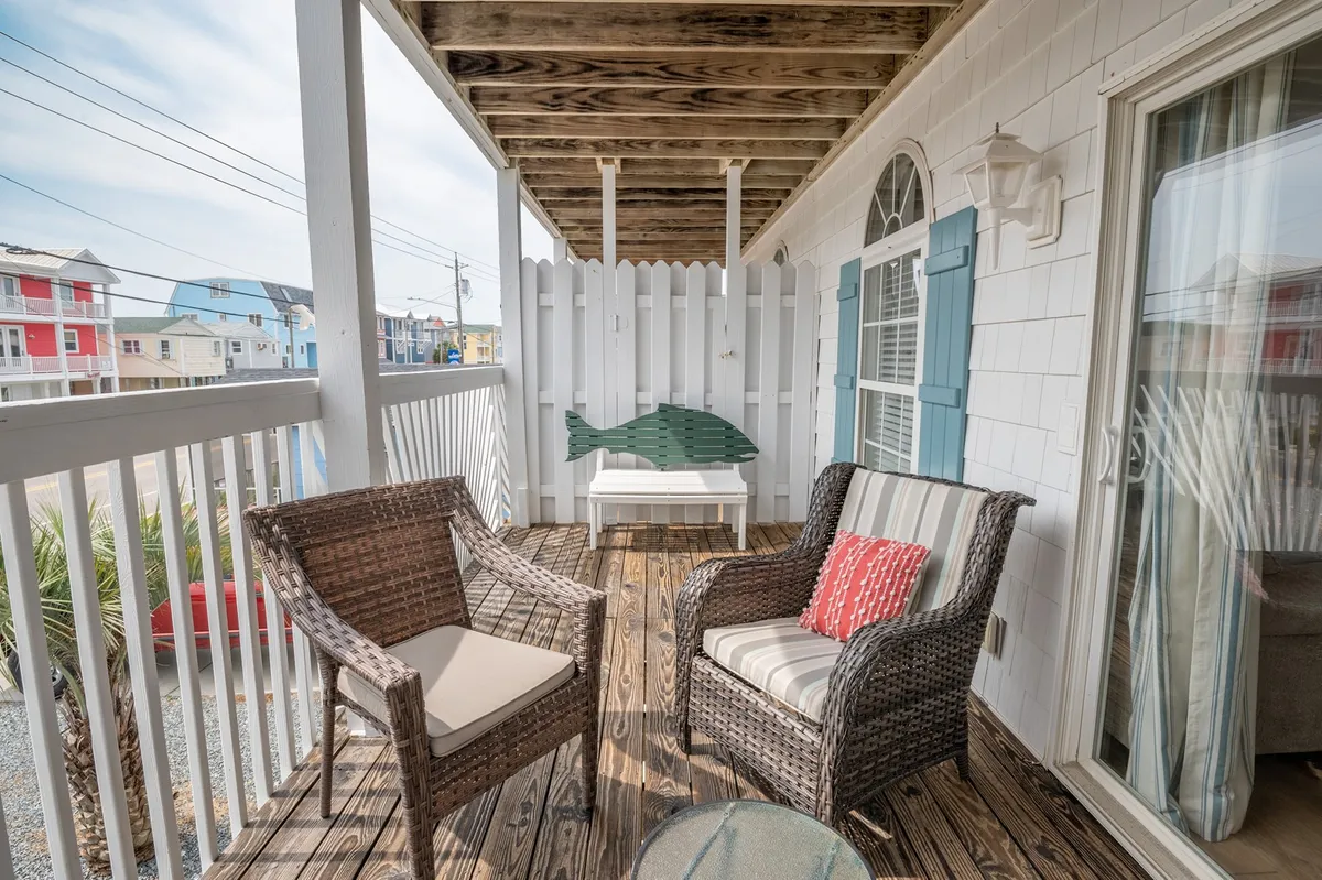 A charming exterior balcony space with wicker chairs, a white table, and blue shutters overlooks a quaint picket fence. It invites guests to sit and soak in the coastal environment, share stories, or plan their next outing.