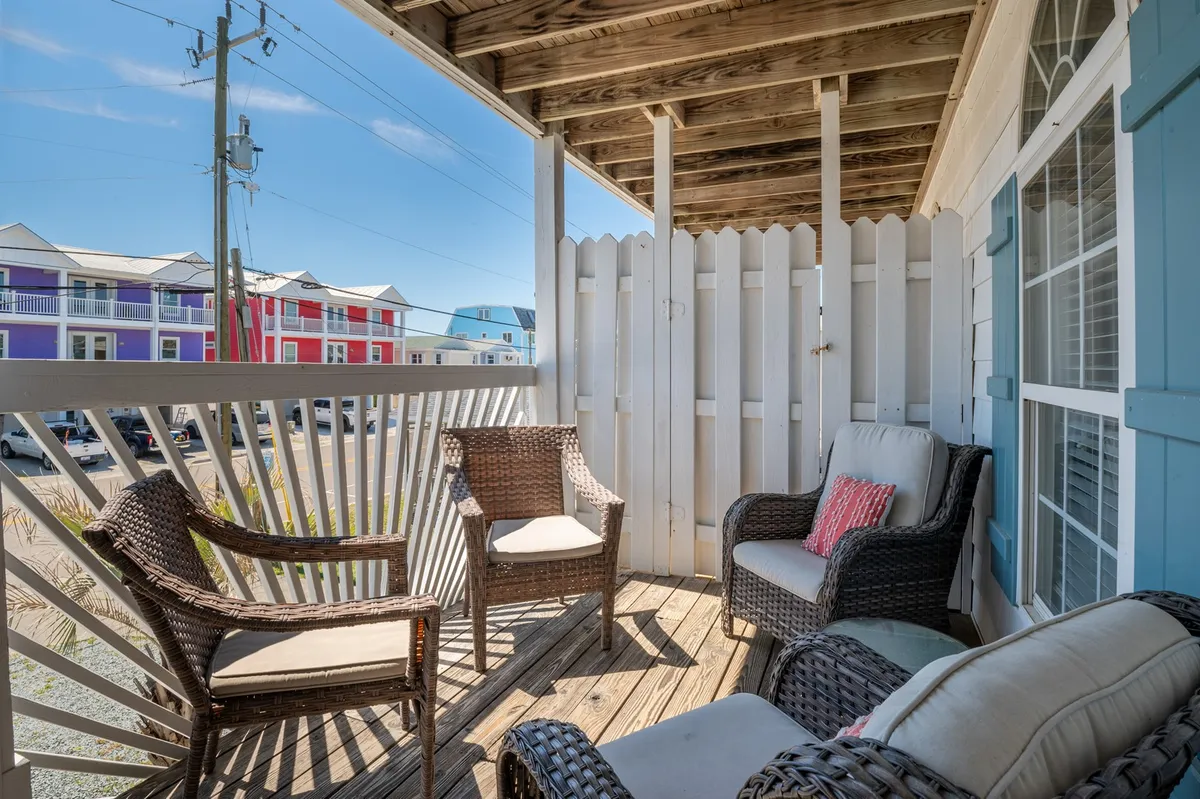 A cozy porch scene featuring wicker chairs and a small table behind a white picket fence. The view suggests a peaceful spot for relaxing, people-watching, or enjoying a sunset, with coastal charm at the forefront.