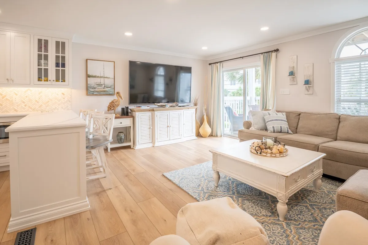 A stylish living room with a comfortable sofa set, modern coffee table, television, kitchen island in the background, and wall artwork. The room is designed for relaxation, entertainment, and seamless socializing, highlighted by a cozy and inviting mood.