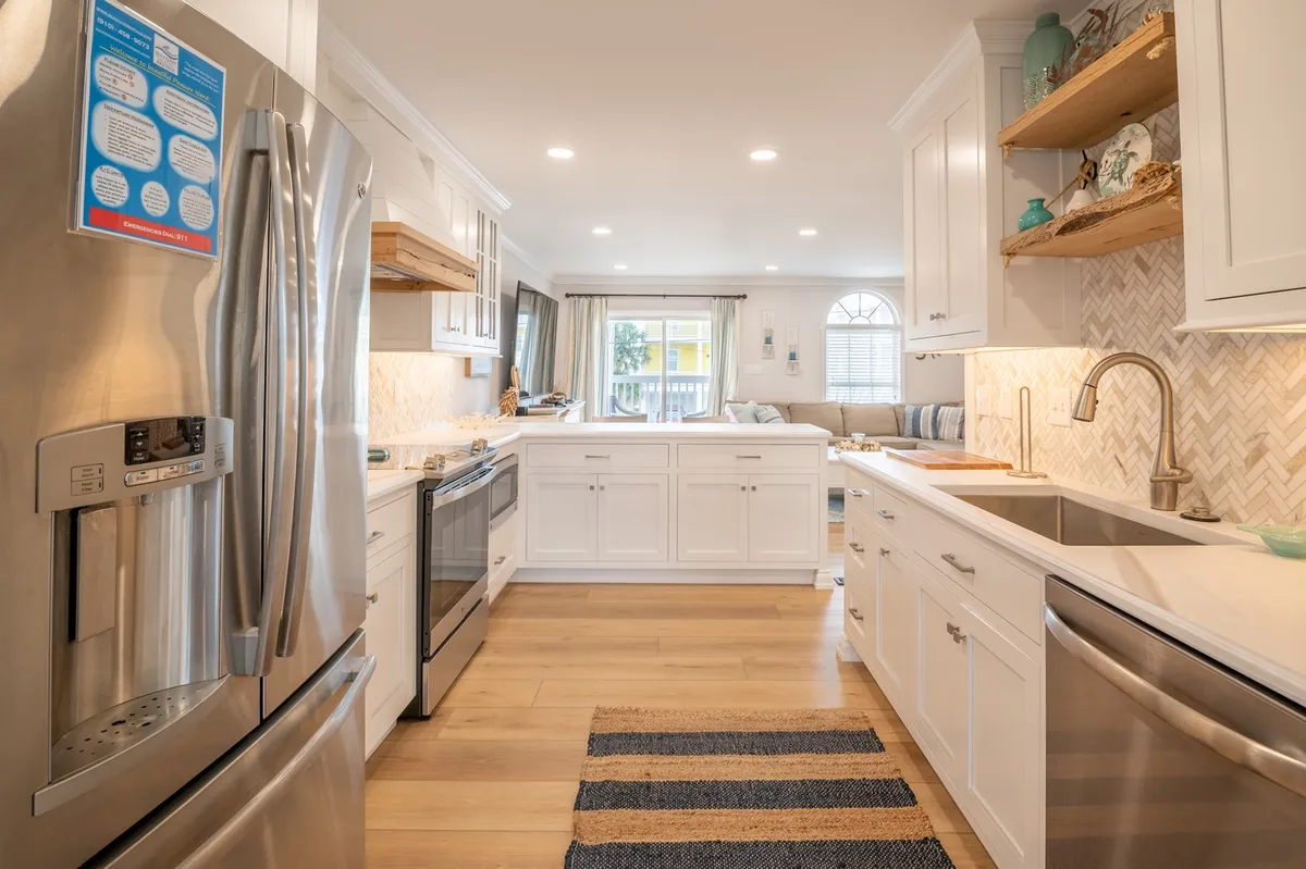 A bright, airy kitchen with gleaming stainless steel appliances, crisp white cabinetry, open shelving, a stylish herringbone backsplash, and a spacious island countertop. The space is inviting and perfectly arranged for cooking, socializing, and family gatherings, with a warm and welcoming atmosphere.
