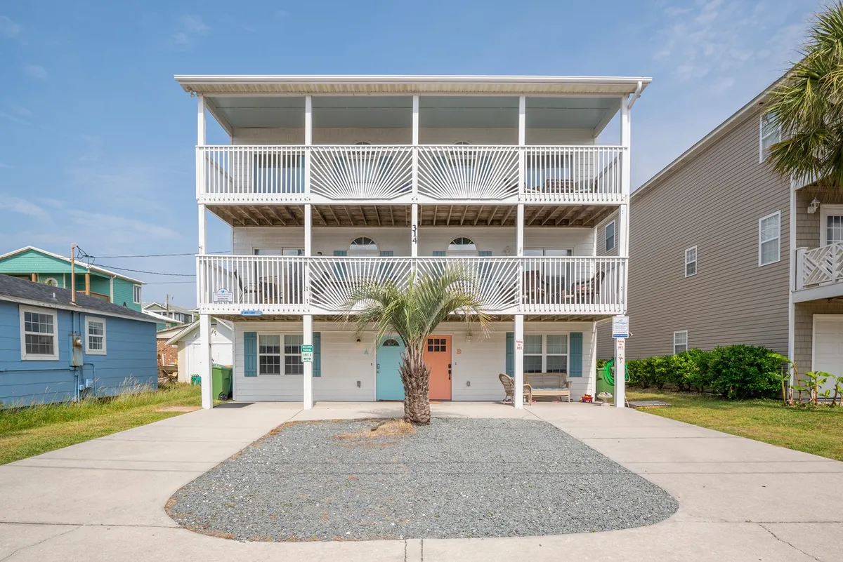 This image shows the vibrant facade of the property, featuring a colorful door, a prominent palm tree, and a cheerful, welcoming porch. The exterior is bathed in natural light, suggesting a lively and inviting atmosphere that fits the mood of a fun beachside destination.