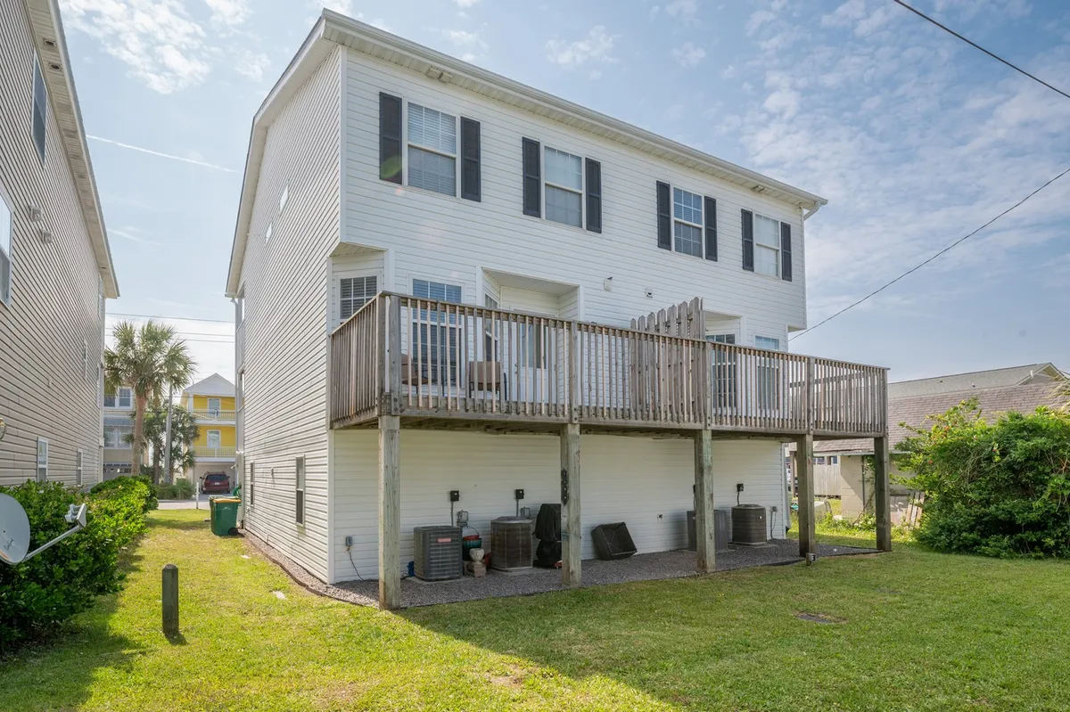 The image showcases the exterior of 'Happy As A Clam,' featuring a spacious deck with white siding, a green lawn, palm trees, and air conditioning units. The setting is ideal for outdoor gatherings, barbecues, or quiet mornings, offering a glimpse of the home's relaxing and communal outdoor environment.
