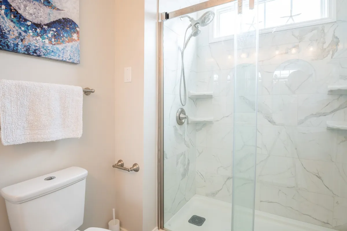 A spa-like ensuite bathroom with a glass shower enclosure, marble shower tiles, a toilet, and tasteful artwork on the wall. The overall effect is serene and luxurious, suggesting a high-end bathing experience.