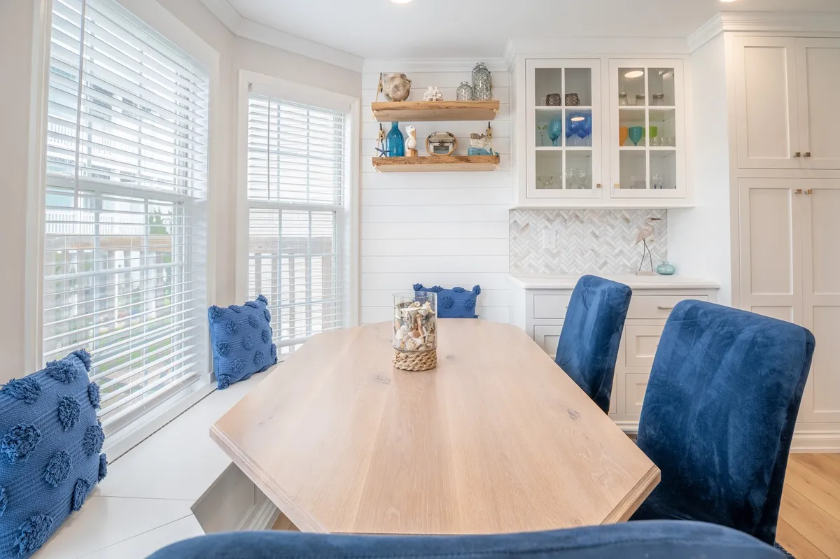 A cozy dining area featuring a wooden table with blue upholstered chairs, shelving with decorative items, and a glass cabinet displaying colorful glassware. The setting is perfect for intimate meals or festive celebrations.