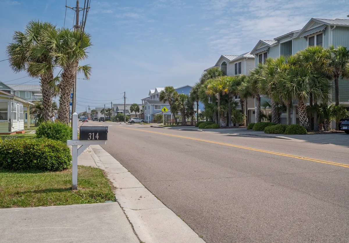 A relaxed street view featuring palm trees, a mailbox marked 314, and a glimpse of nearby houses. The scene invites viewers to imagine themselves strolling or biking through the beach neighborhood, soaking up the laid-back coastal vibe.