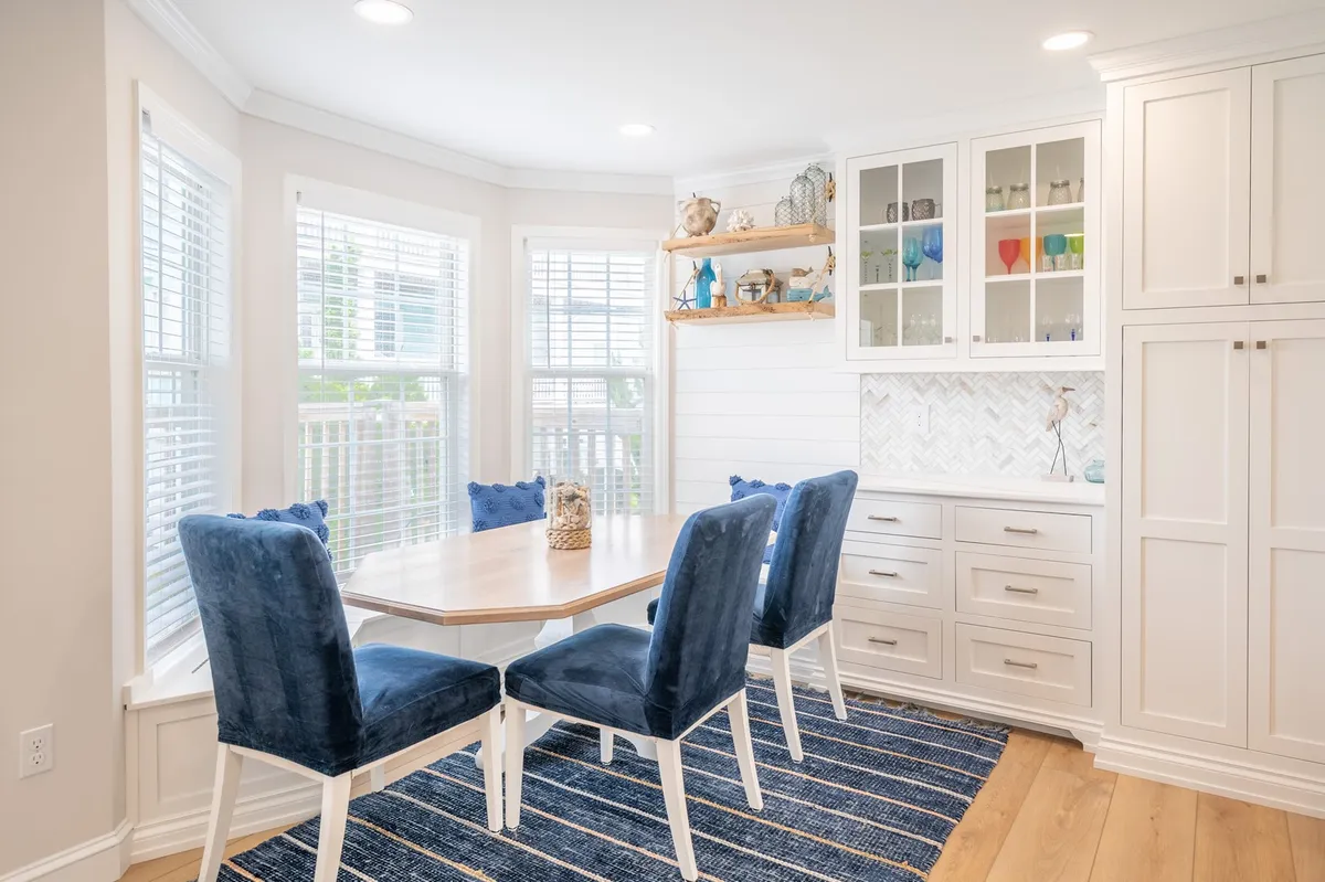 A spacious dining area with a large table surrounded by plush blue velvet chairs, flanked by a glass cabinet filled with colorful items and decorative shelving. The setting is warm, welcoming, and perfect for both family meals and festive gatherings.