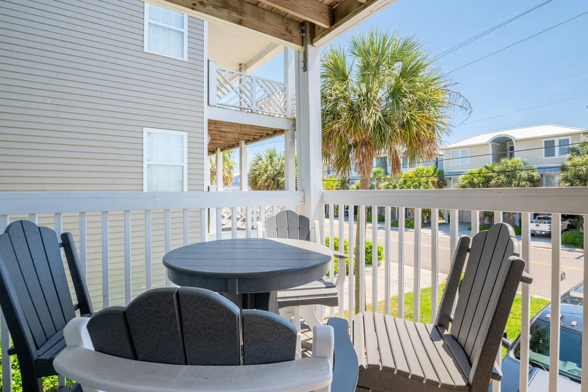 An exterior view of an inviting outdoor seating area, featuring comfortable chairs and a table shaded by palm trees. The vantage point provides a glimpse of the neighborhood street, evoking a casual, friendly atmosphere ideal for people-watching or relaxing between beach adventures.