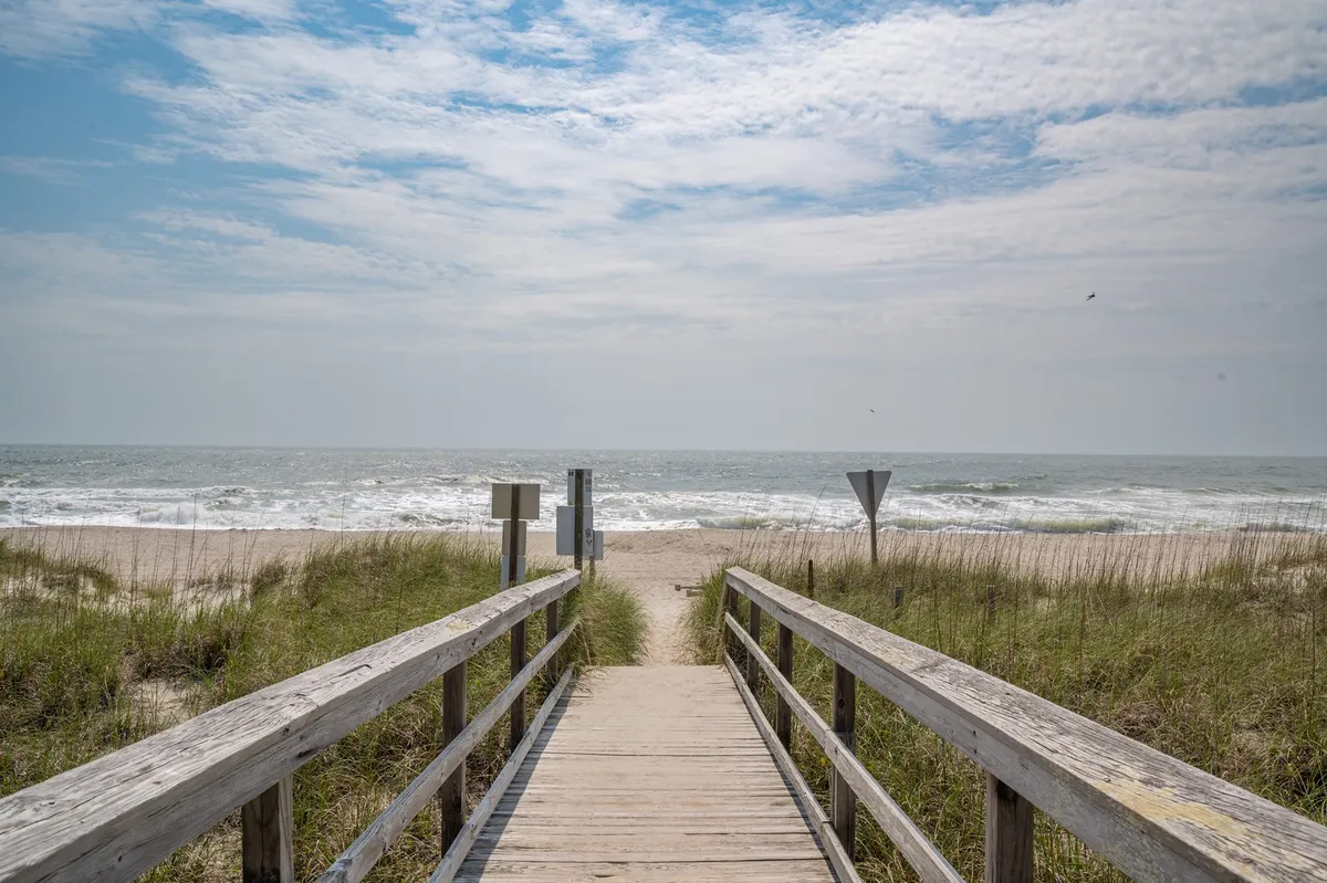 This image captures a tranquil wooden boardwalk stretching over dune grass, leading directly to the sandy shoreline. Gentle ocean waves roll onto the sand as the open sky provides a serene backdrop. The boardwalk invites viewers to imagine walking straight to the beach.