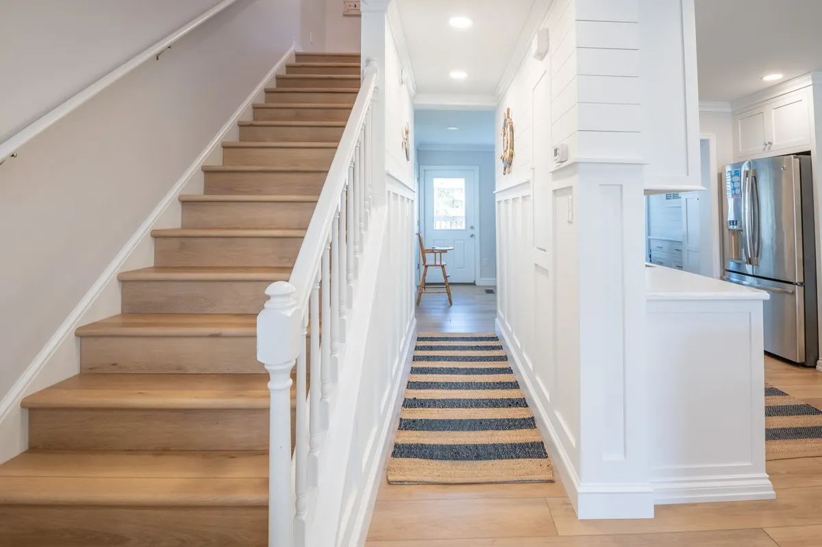 This image shows a bright, welcoming entryway with a visible staircase leading up and a glimpse of the kitchen area beyond. The striped rug and open sightlines suggest ease of movement between levels and gathering spaces.