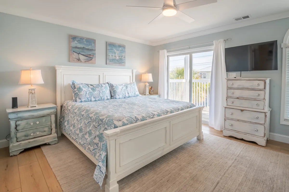 A tranquil bedroom featuring a bed with patterned bedding, a nightstand with a lamp, a large window allowing in natural light, and a dresser along one wall. The room emanates a serene, cozy vibe and feels welcoming for restful nights or calm morning routines.