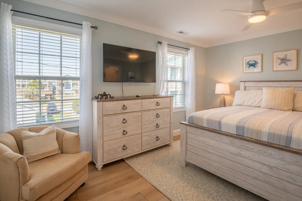 A cozy bedroom with a comfortable bed, dresser, armchair, TV, and framed art on the wall. Natural light filters in, creating a peaceful retreat ideal for relaxing, watching TV, or enjoying quiet moments.