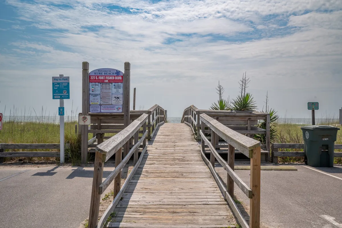 A wooden walkway leads past lush greenery toward a beach access sign, with the ocean just beyond and parking signage visible. The setting sun casts a golden glow, emphasizing the proximity to sand, surf, and classic seaside activities.